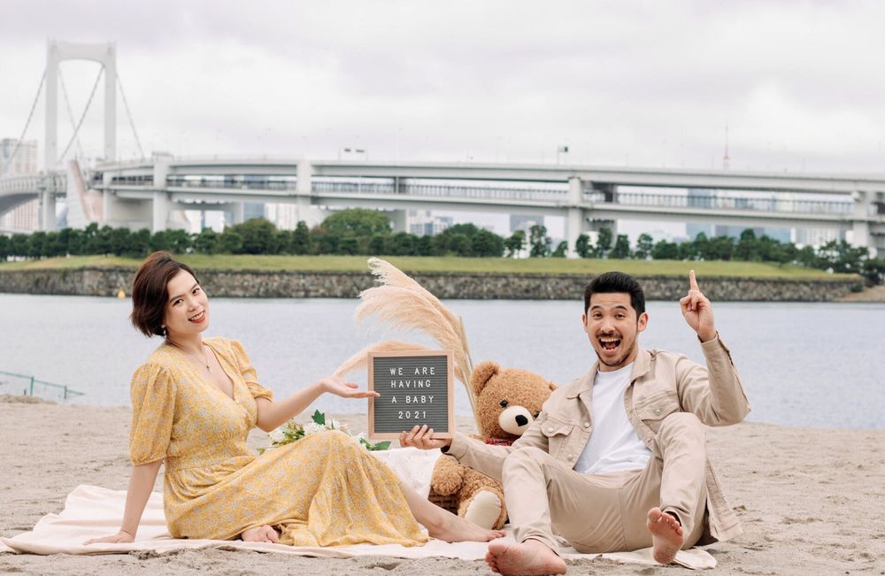 Couple having picnic with teddy bear near Rainbow Bridge, holding celebration sign.