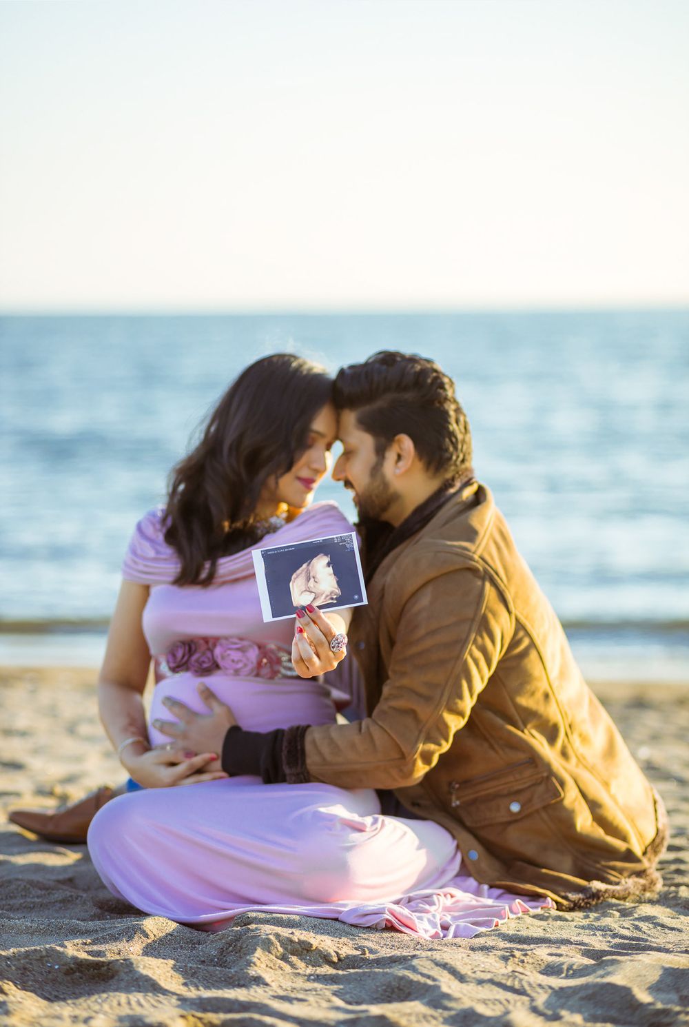 Couple sitting on beach sand holding ultrasound photo during sunset maternity photography session.