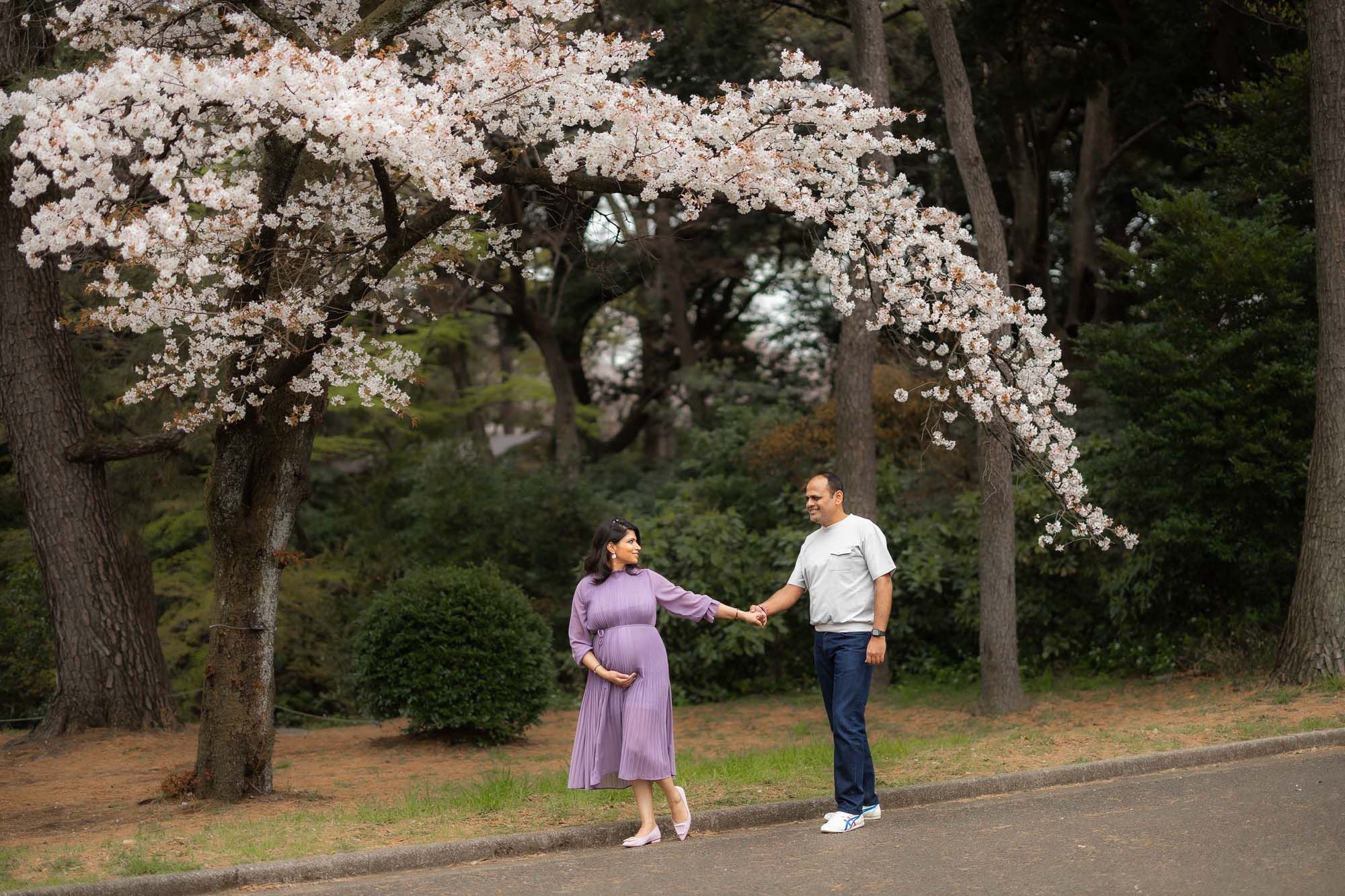 Couple holds hands under blooming cherry blossom trees during spring maternity photo session.