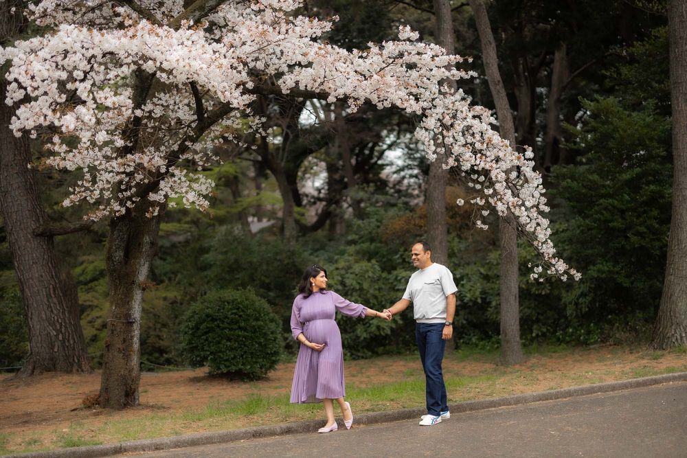 Couple holds hands under blooming cherry blossom trees during spring maternity photo session.