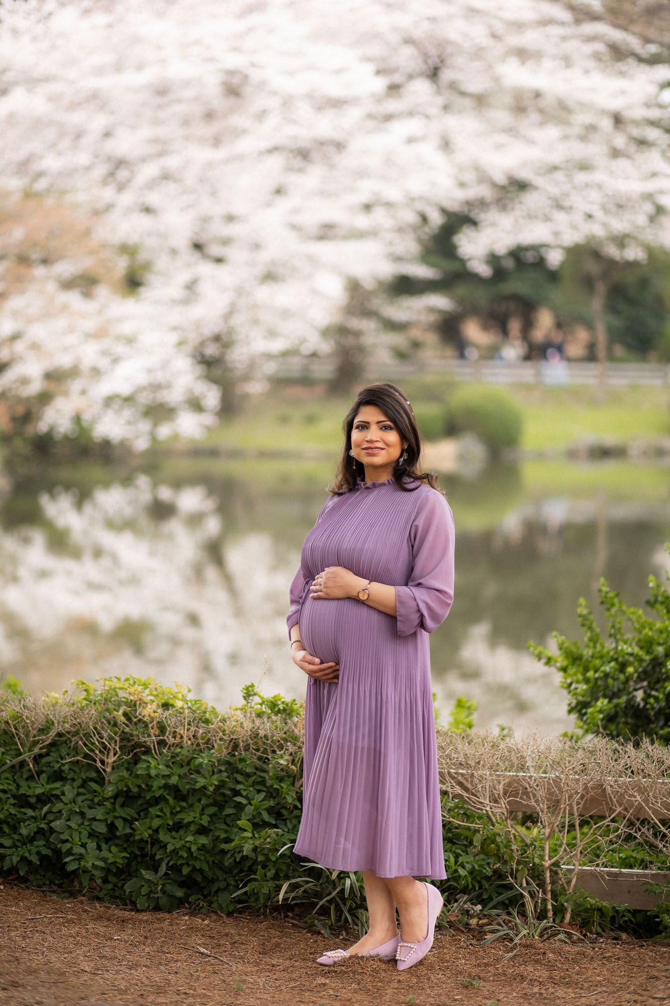 Pregnant woman poses by lake with cherry blossoms in full bloom during spring photo session.