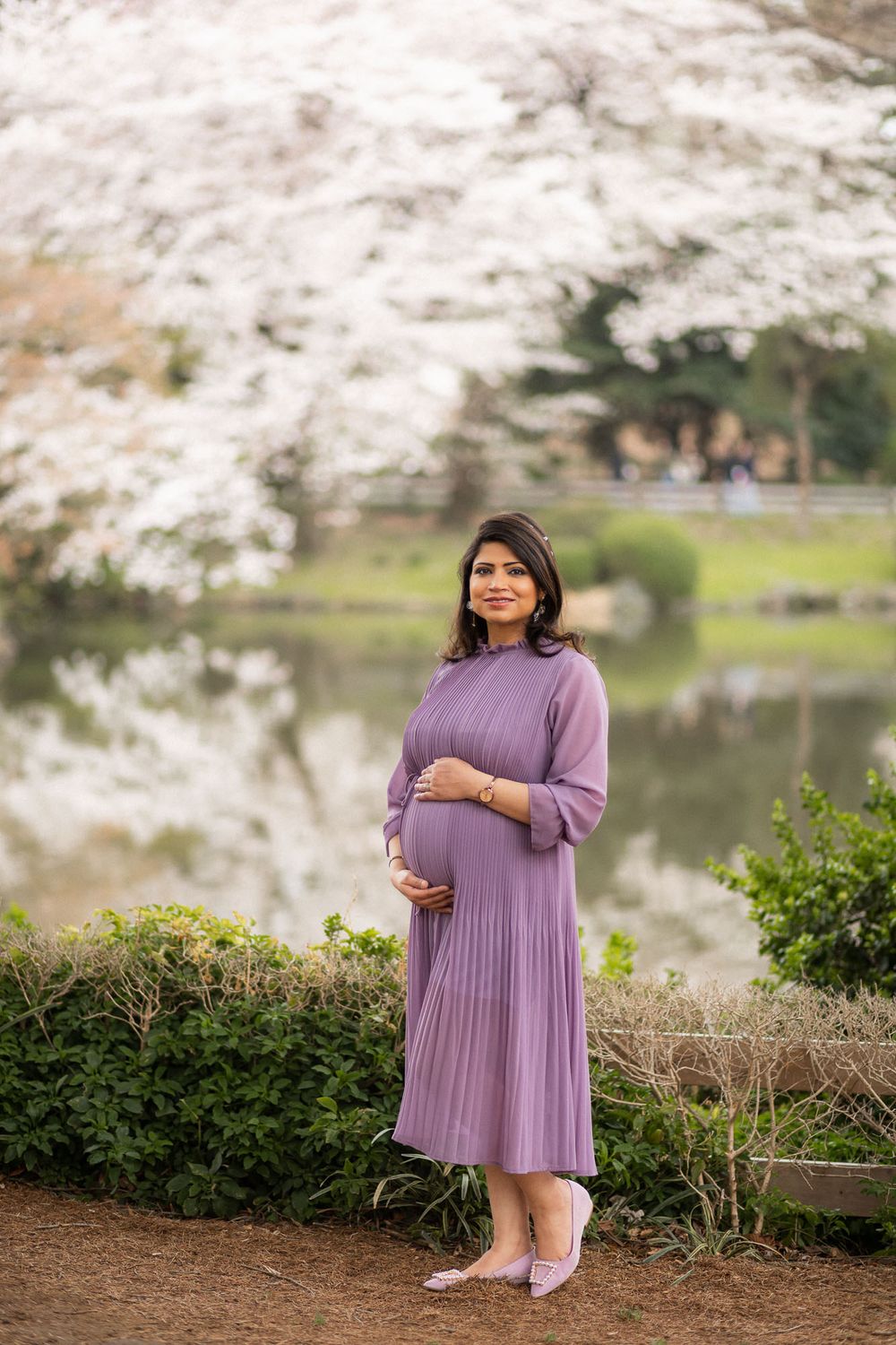 Pregnant woman poses by lake with cherry blossoms in full bloom during spring photo session.