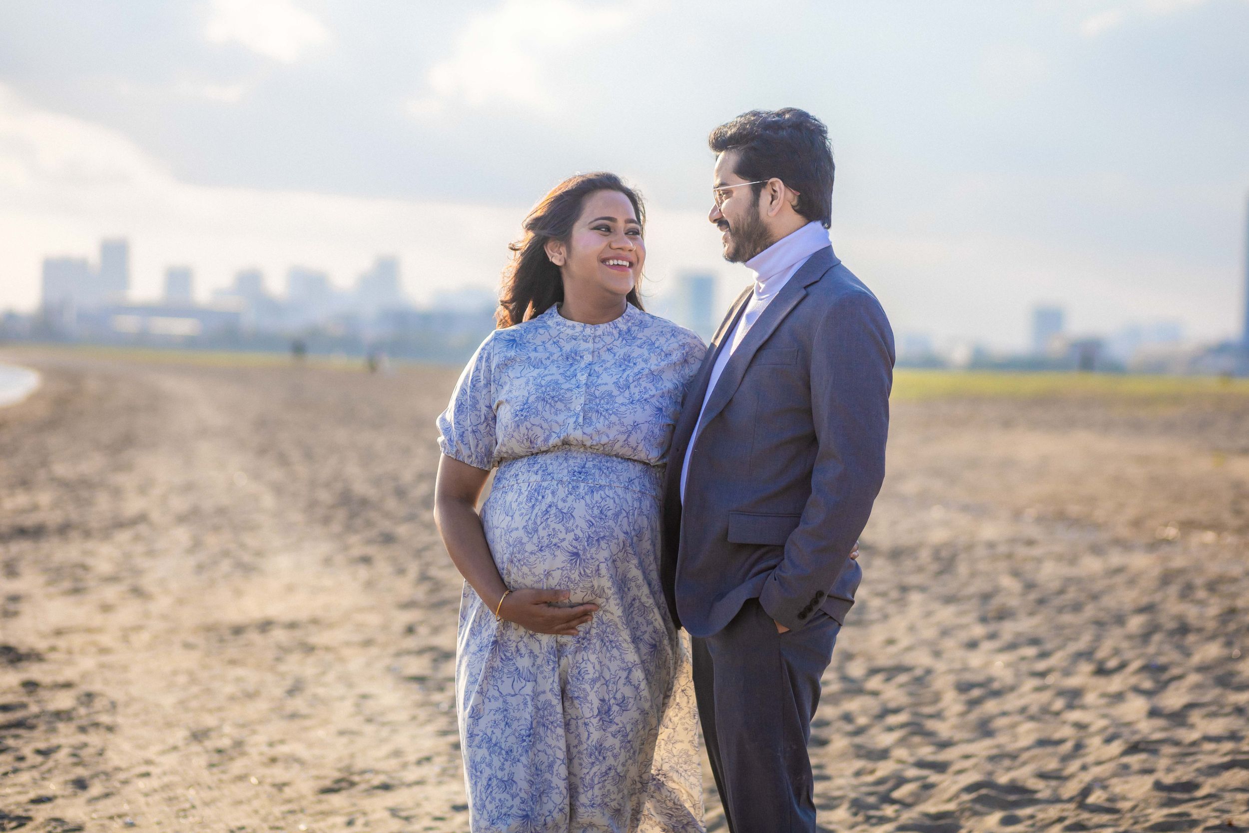 Couple poses on beach with city skyline visible in background.