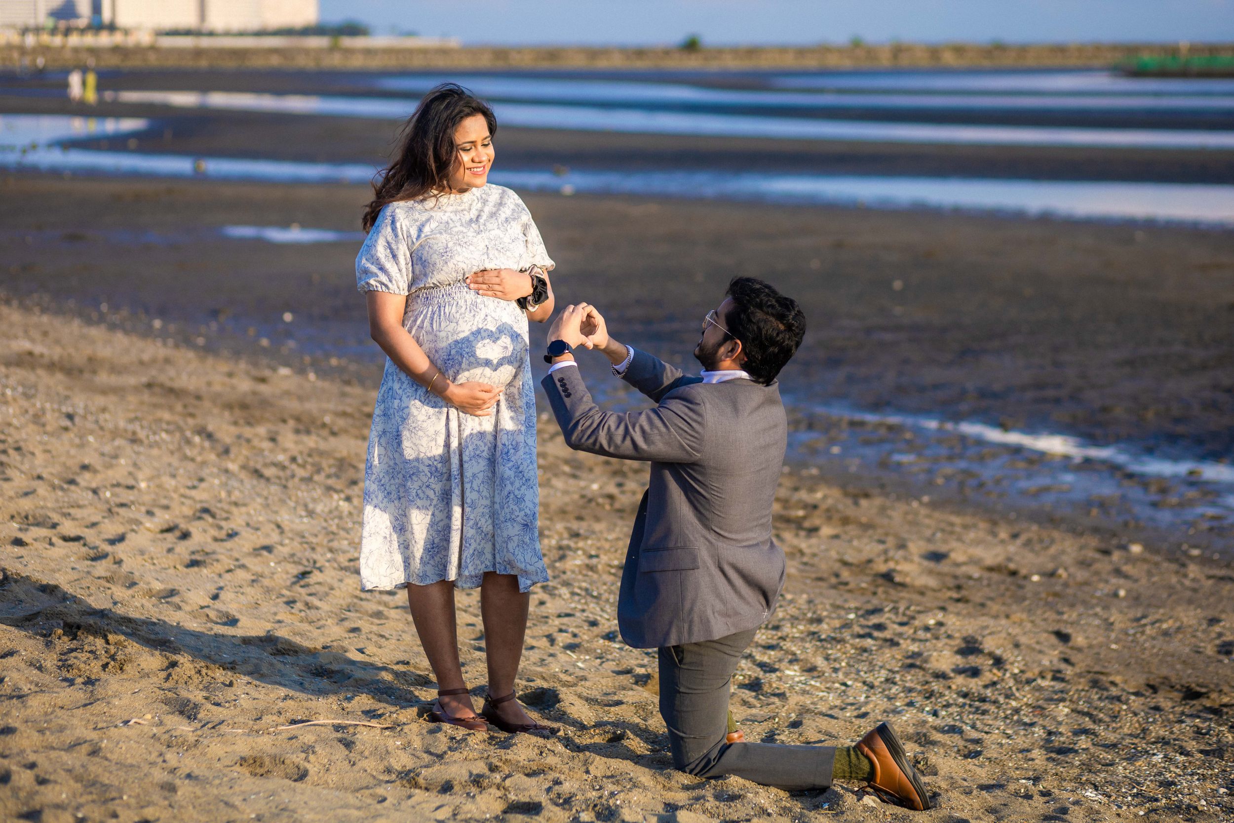 A romantic proposal moment takes place on a sandy beach during sunset with calm water in the background.