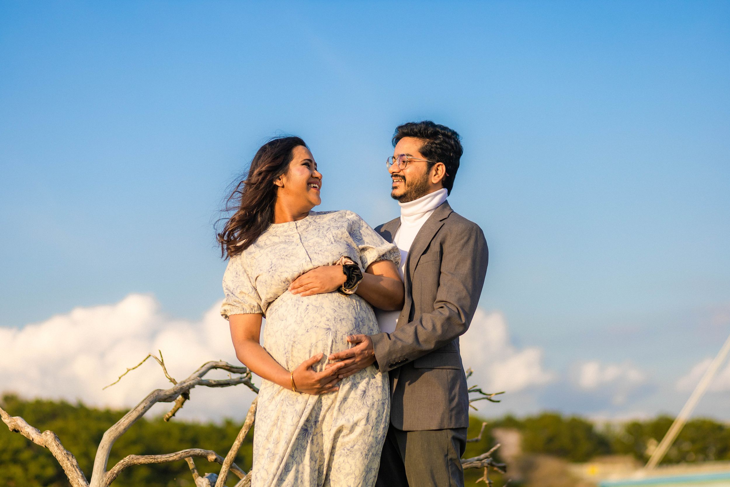 A maternity photoshoot featuring a couple embracing against a bright blue sky with white clouds.
