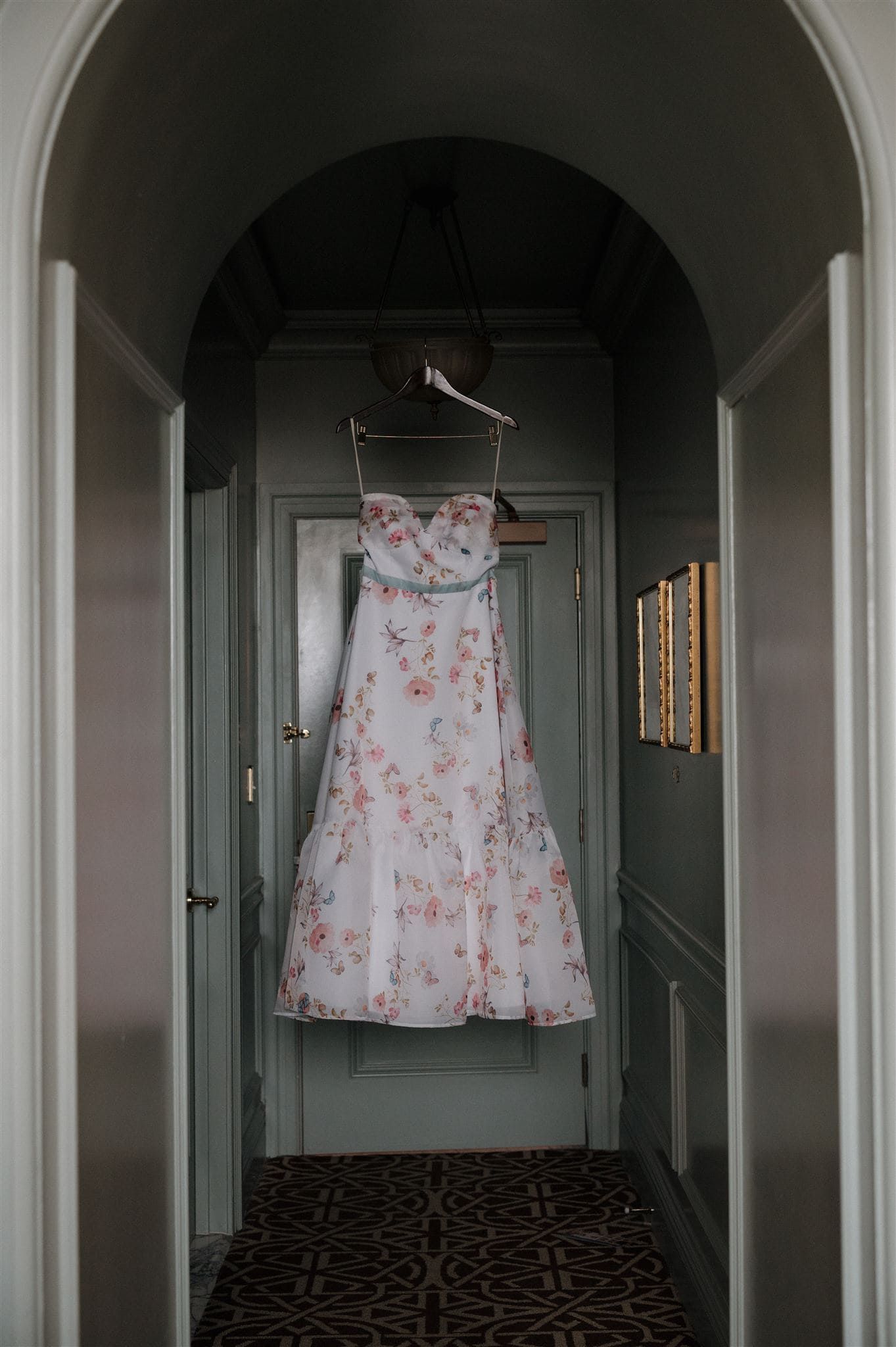 Floral wedding dress hanging in arched hallway with gray-green walls and patterned carpet.