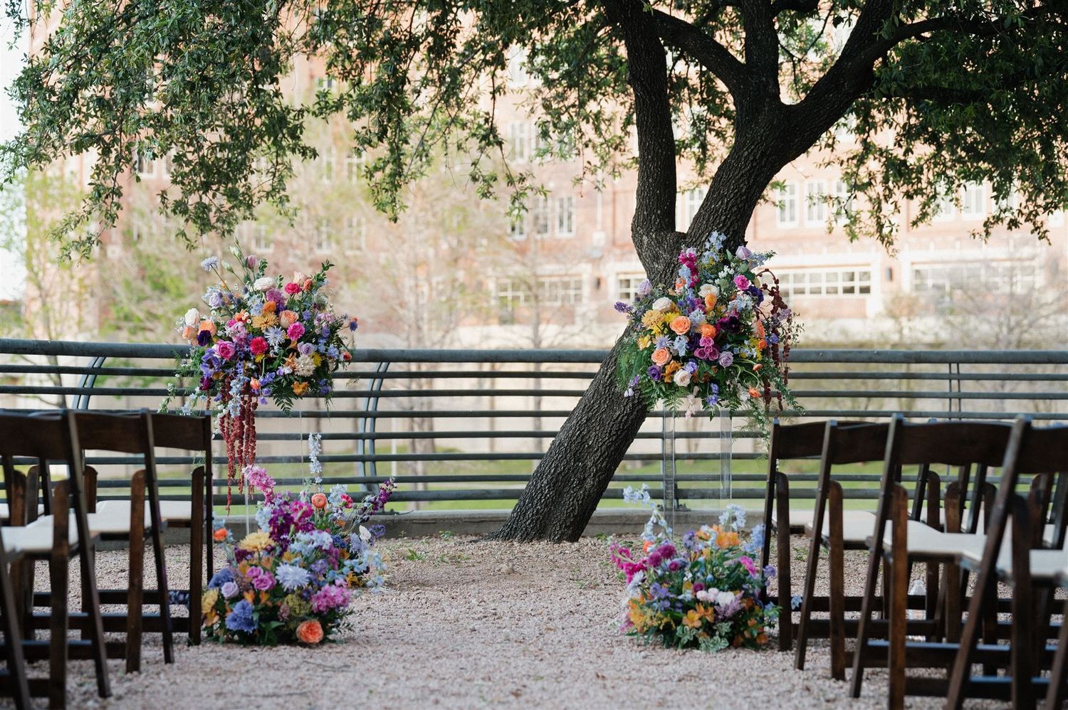 Outdoor wedding ceremony setup with wooden chairs and colorful floral arrangements under a tree.