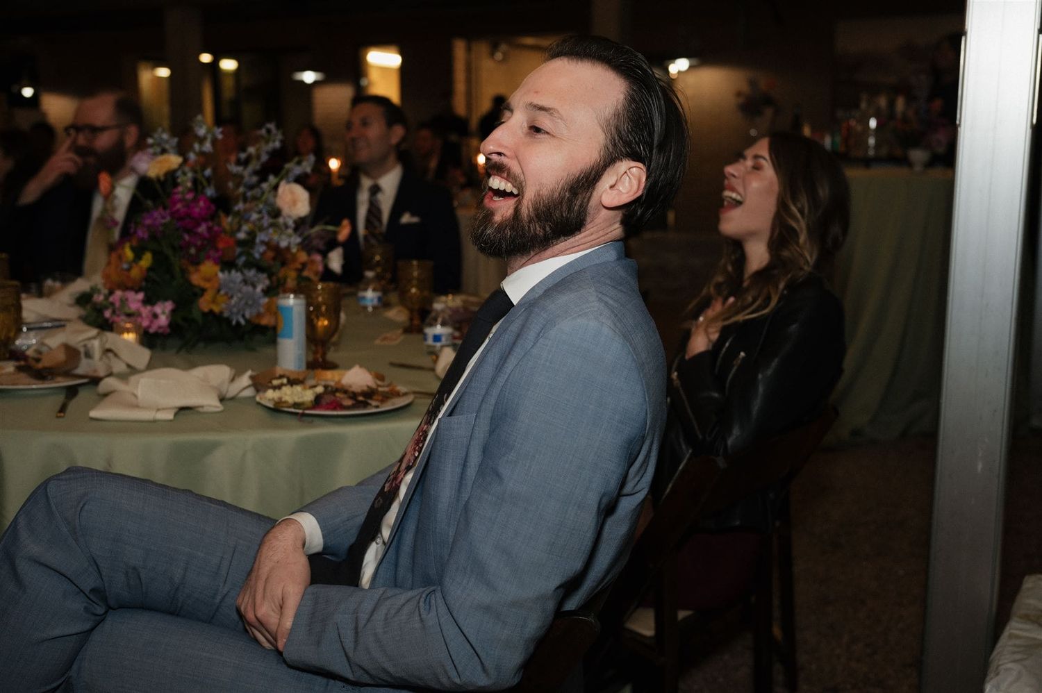 Guest in gray blazer laughs during an evening dinner party with tables and decorations visible in background.
