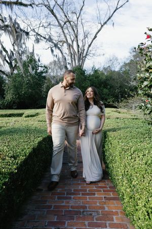 Pregnant couple walking hand-in-hand through a lush garden maze on a brick path, sharing a loving glance.