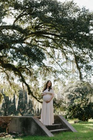 Pregnant woman in blush gown stands under sprawling oak tree with Spanish moss on stone steps outdoors.