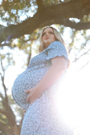 A pregnant woman in a floral dress stands in sunlit nature during a maternity photoshoot.