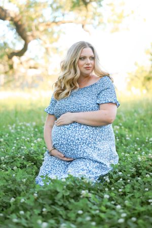Expectant mother in blue floral dress poses gracefully in a blooming white flower field during golden hour.