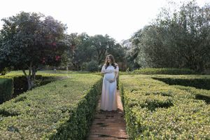 Pregnant woman in white dress standing in lush green garden maze pathway surrounded by trimmed hedges and trees.