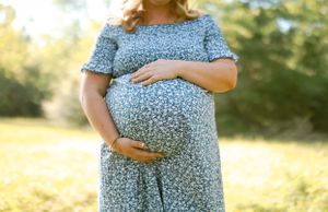 A pregnant woman in a floral blue dress stands in a sunlit field during a golden hour maternity photoshoot.