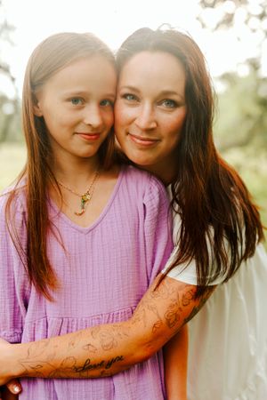 A mother and daughter embrace while wearing purple and white tops in golden sunset lighting with henna tattoo visible.
