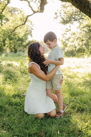 A mother kneels in a sunlit green field, tenderly embracing her young son beneath a large oak tree.