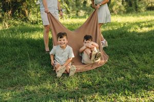 Two young boys sit on a blanket held by adults in a sunny green park during a family outdoor photo session.