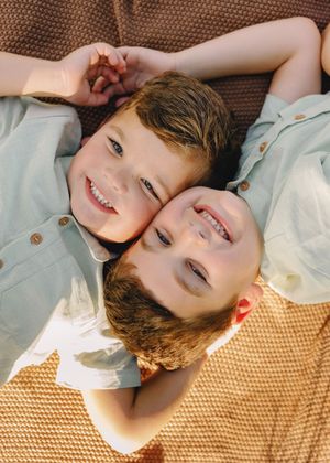 Two smiling young boys lying head-to-head on a woven blanket, wearing light blue shirts outdoors.