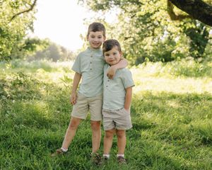 Two young brothers in matching sage green shirts and khaki shorts pose together in a sunny green meadow.