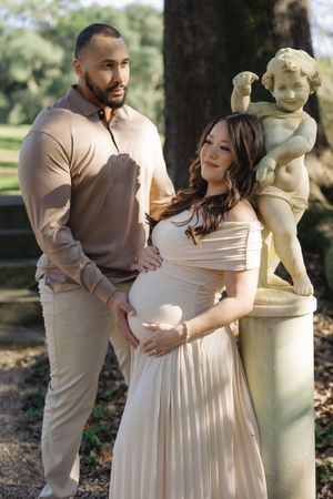 Couple poses for maternity photo shoot outdoors near cherub statue, woman in cream dress cradling baby bump.
