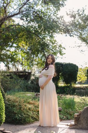Pregnant woman in elegant off-shoulder blush gown cradling baby bump in lush garden setting.