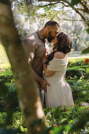 Couple sharing a romantic kiss in a lush garden, man in tan shirt and woman in off-shoulder white dress.