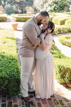 Couple in neutral tones embracing in a garden, pregnant woman in blush maxi dress smiling up at partner.