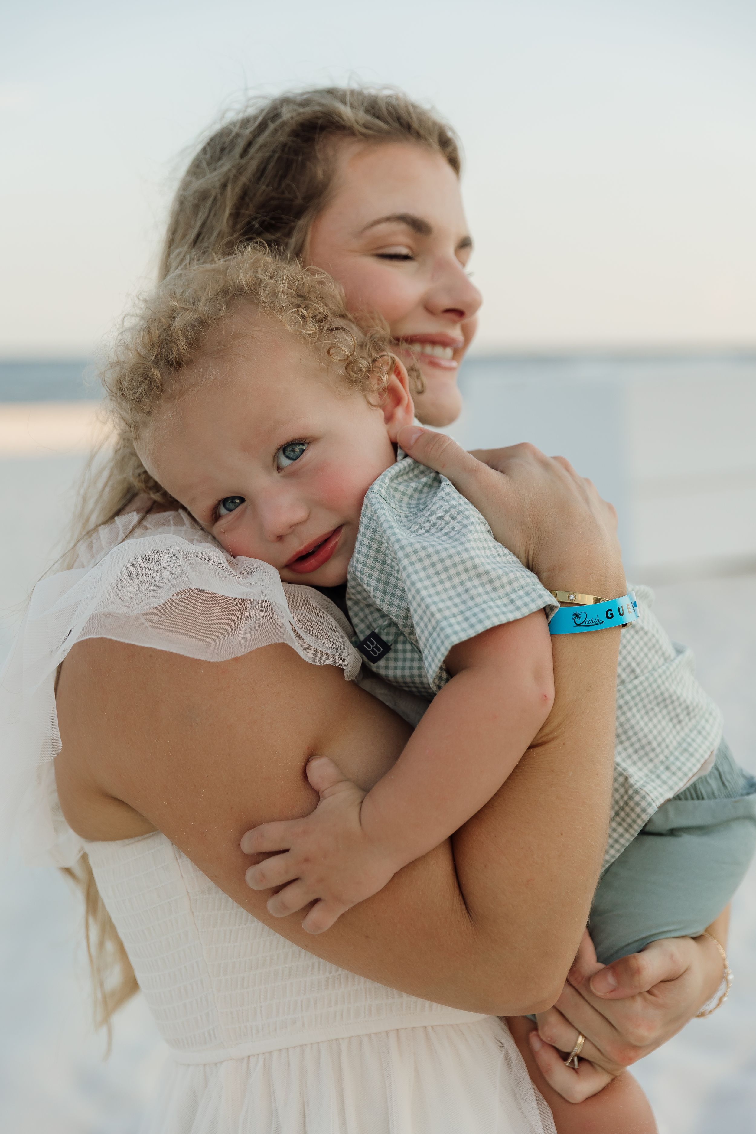 A smiling mother in a white dress tenderly holds her curly-haired toddler boy near a beach at sunset.