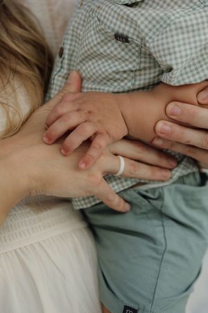 A mother wearing a white ring holds her young child's hands, dressed in a green plaid shirt and sage shorts.