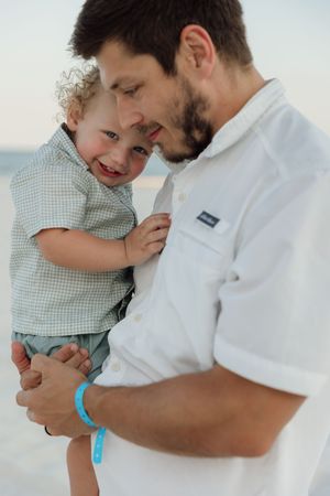 A bearded man in a white shirt holds a smiling curly-haired toddler boy on a beach.