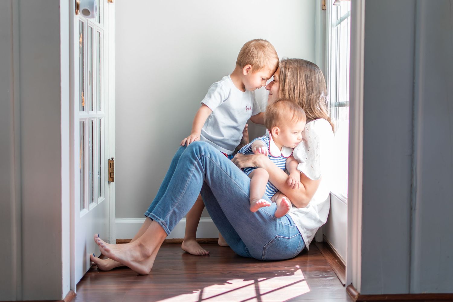 A mom and her two sons share a tender moment snuggled on the floor in front of the back door with light pouring in.