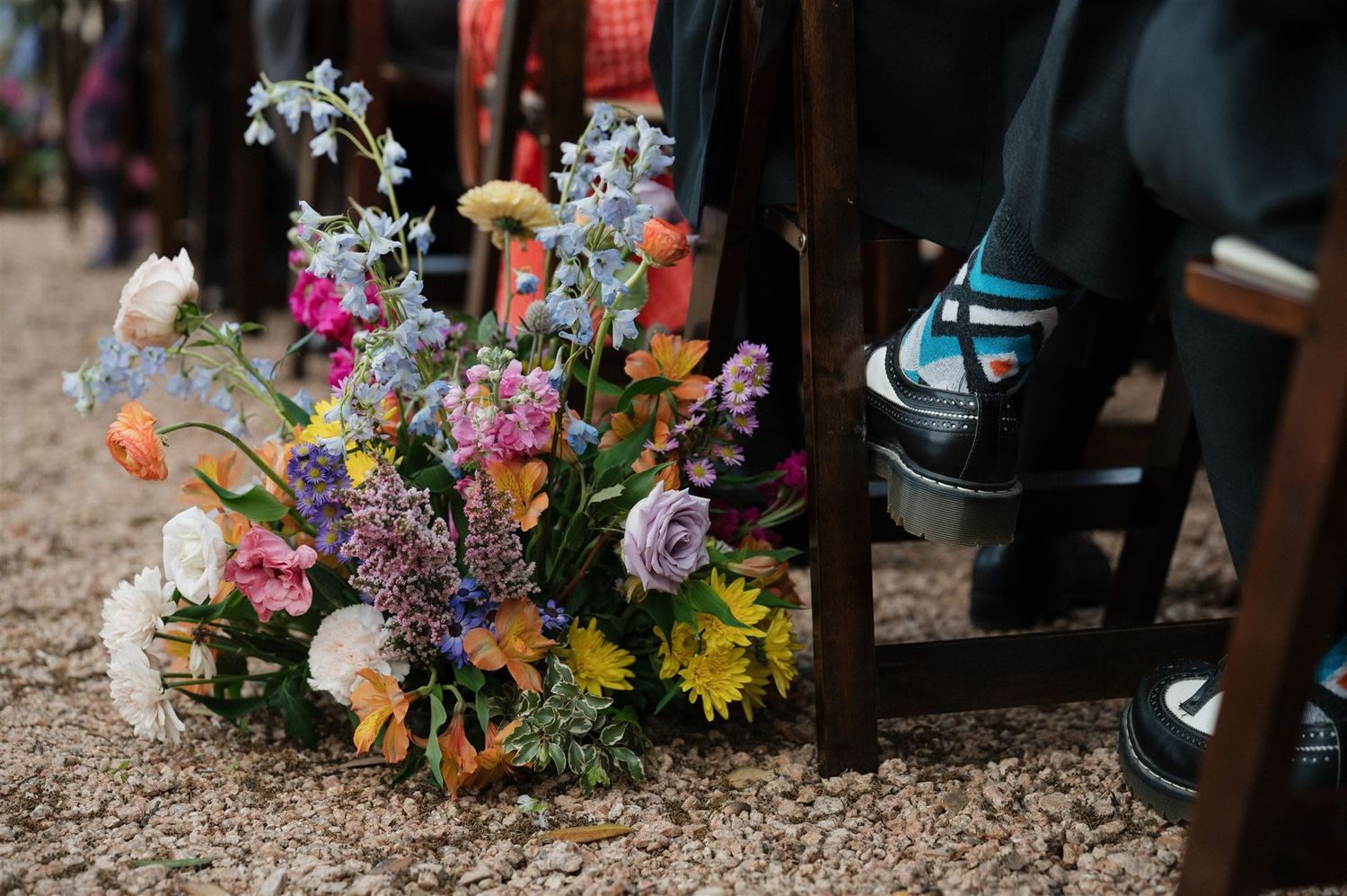 Vibrant wildflower bouquet with daisies and purple blooms sits on ground next to black dress shoes.