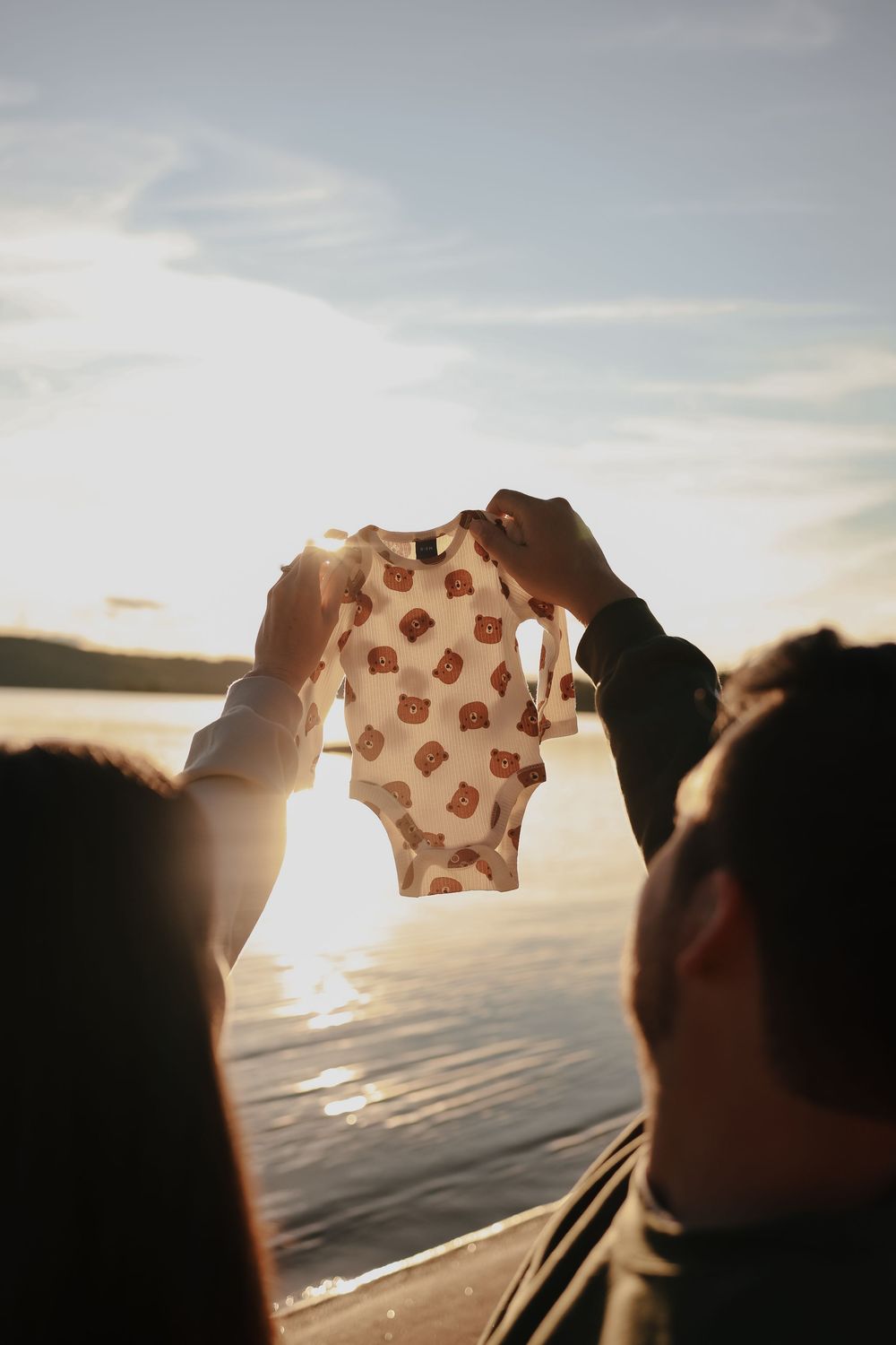 A small baby onesie with a pattern is held up against a sunset sky over water, creating a silhouetted scene.