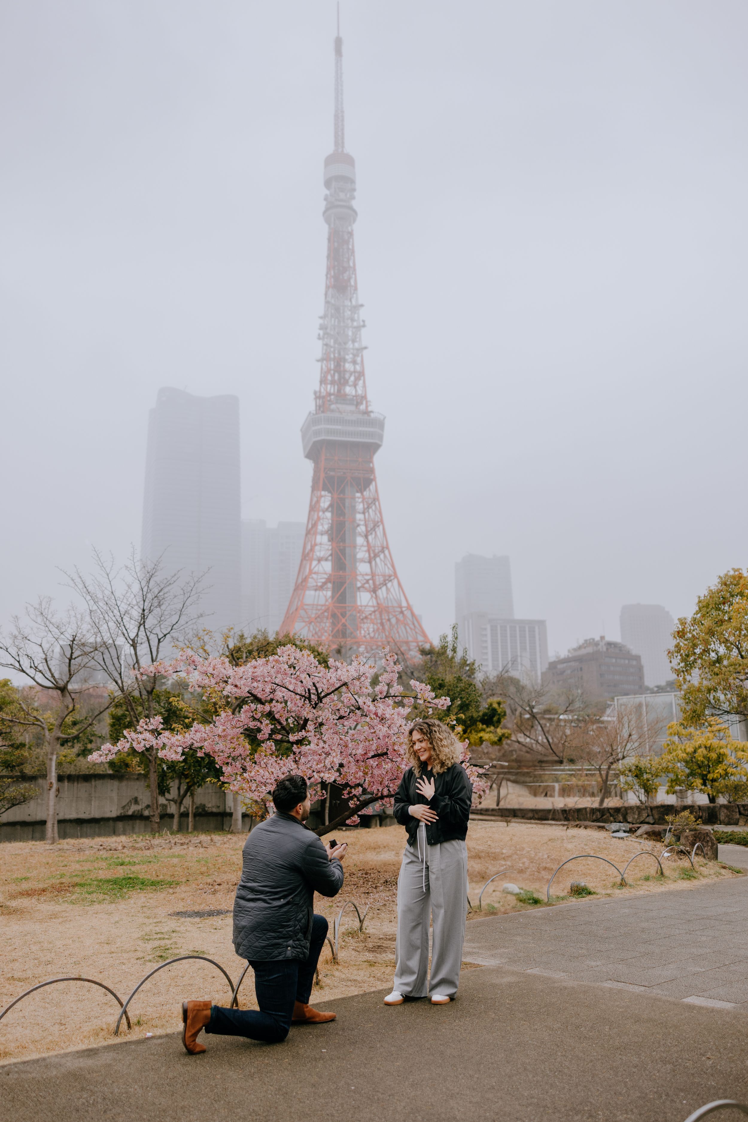 A romantic proposal scene in front of Tokyo Tower during cherry blossom season on a foggy spring day.