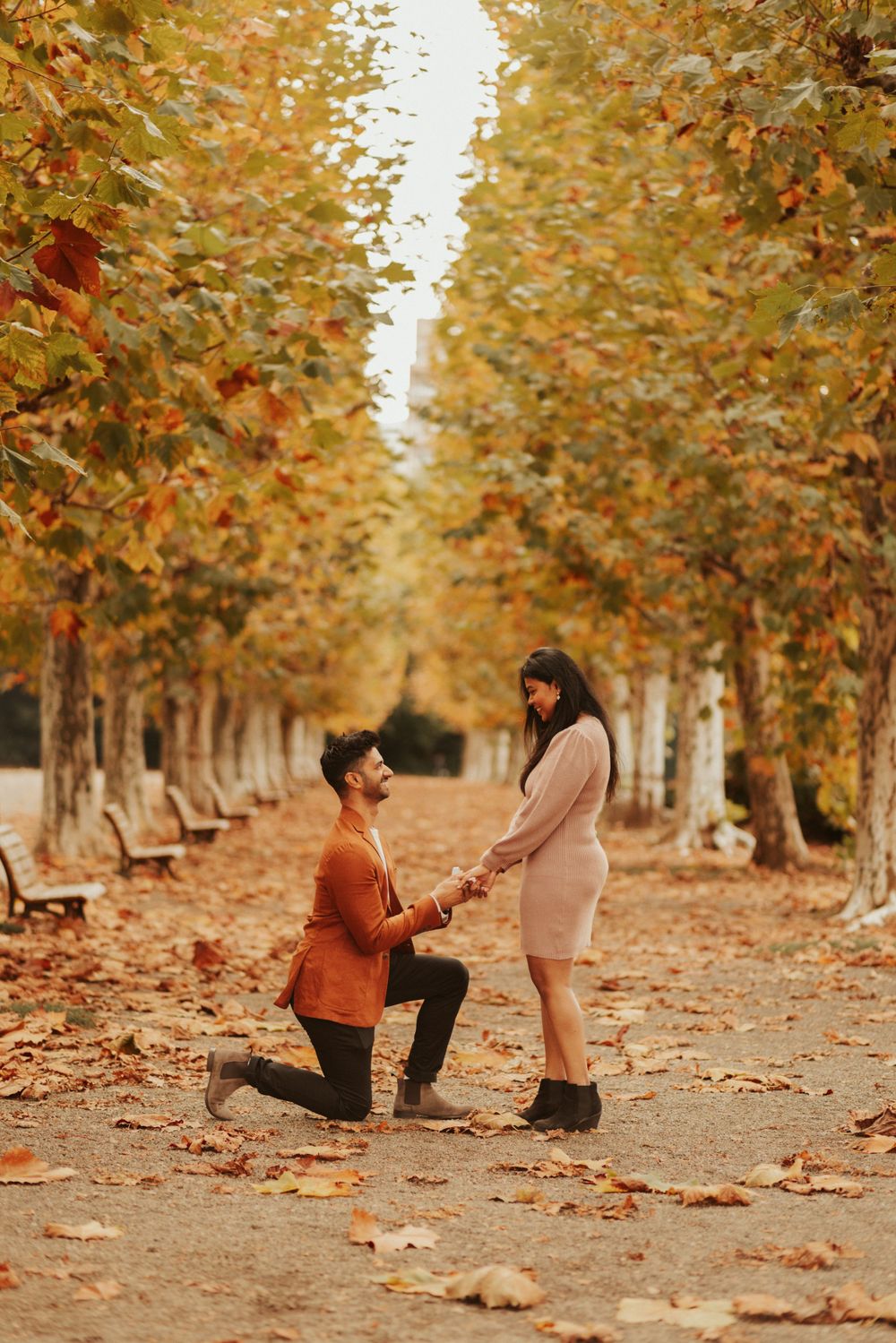 A romantic autumn proposal scene between two people on a tree-lined path covered in fallen leaves.