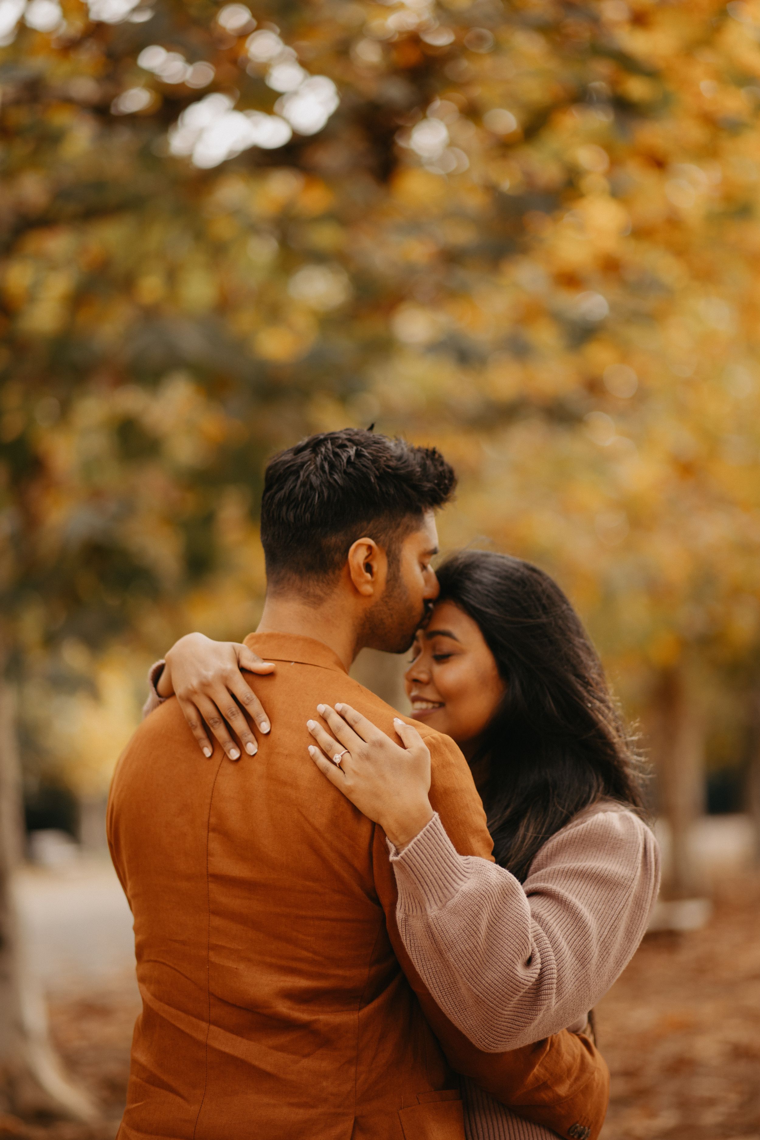 A couple embraces tenderly in a park during autumn, surrounded by golden fall foliage.
