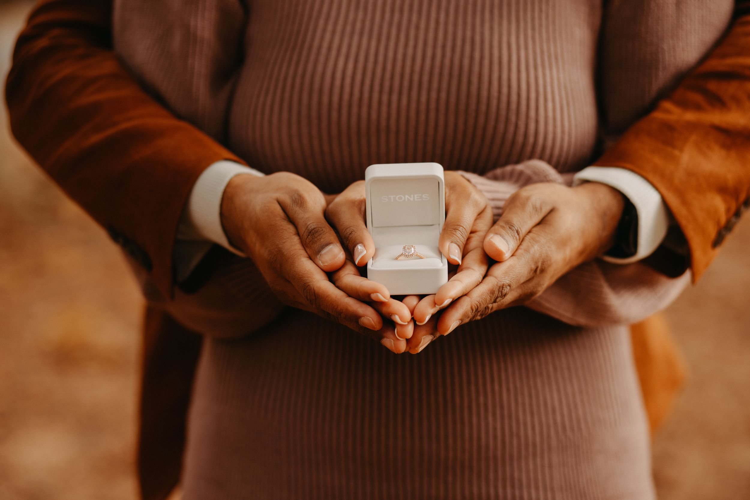 Hands holding a small white ring box against warm autumn-colored clothing for an intimate engagement moment.