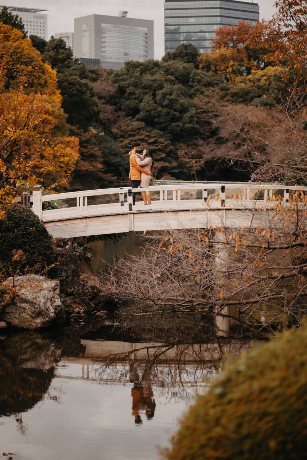 A couple embraces on a white bridge over a tranquil pond surrounded by fall foliage and urban buildings.