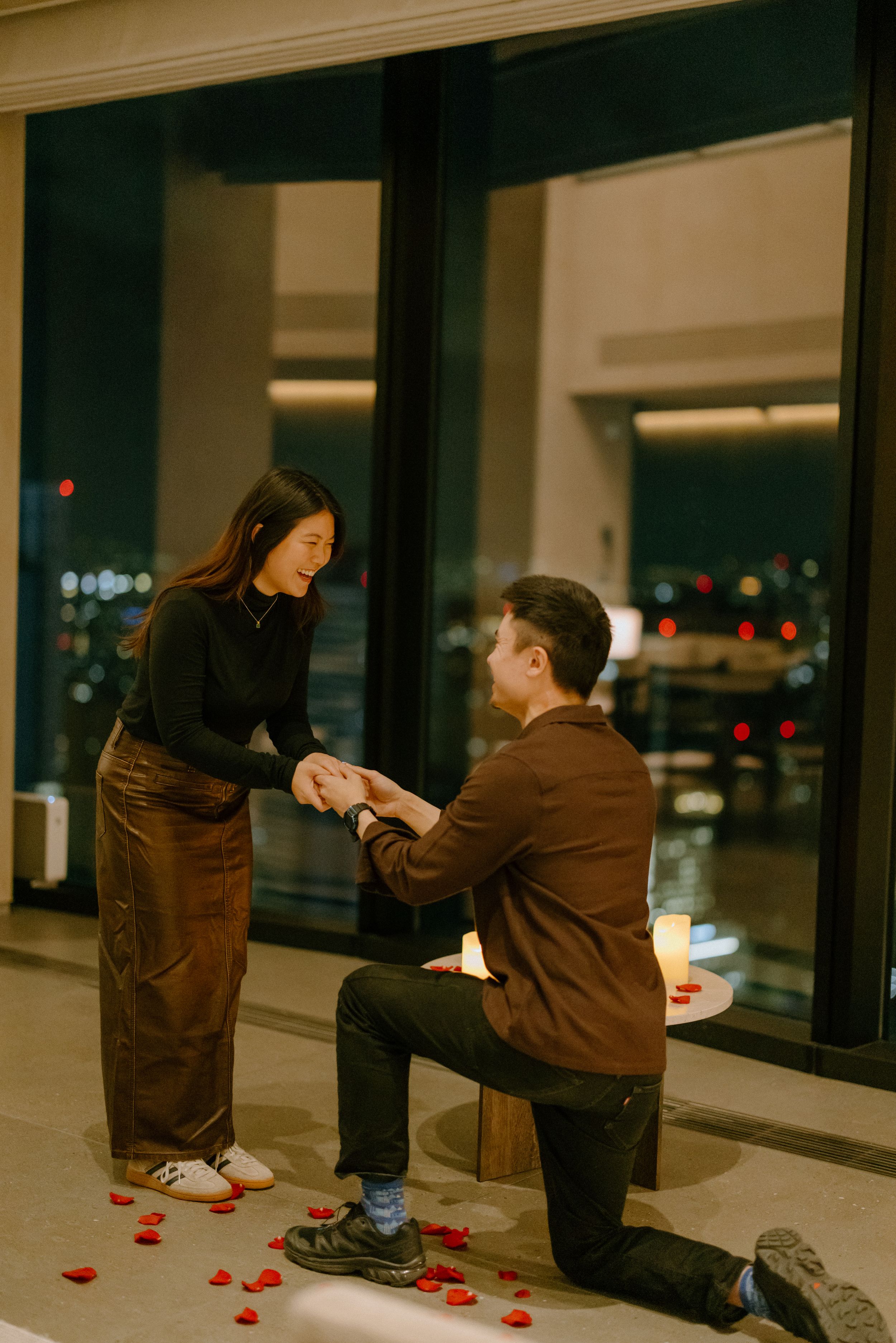 A romantic proposal scene at night with rose petals scattered on the floor by large windows overlooking city lights.