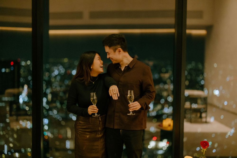 Romantic couple sharing intimate moment while holding wine glasses against city skyline view at night through windows.