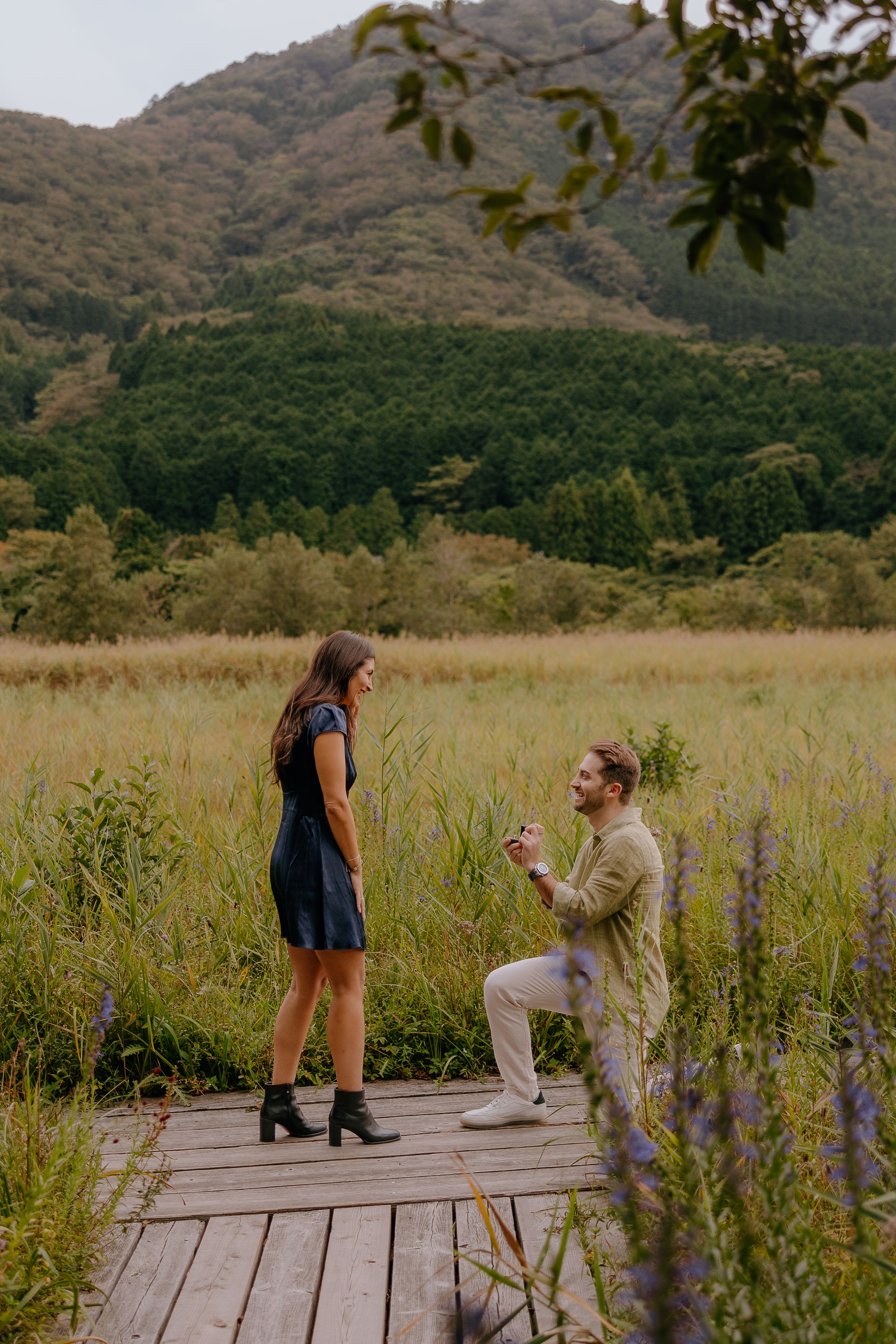 A couple shares a special moment on a wooden deck surrounded by tall grass and mountains in the background.