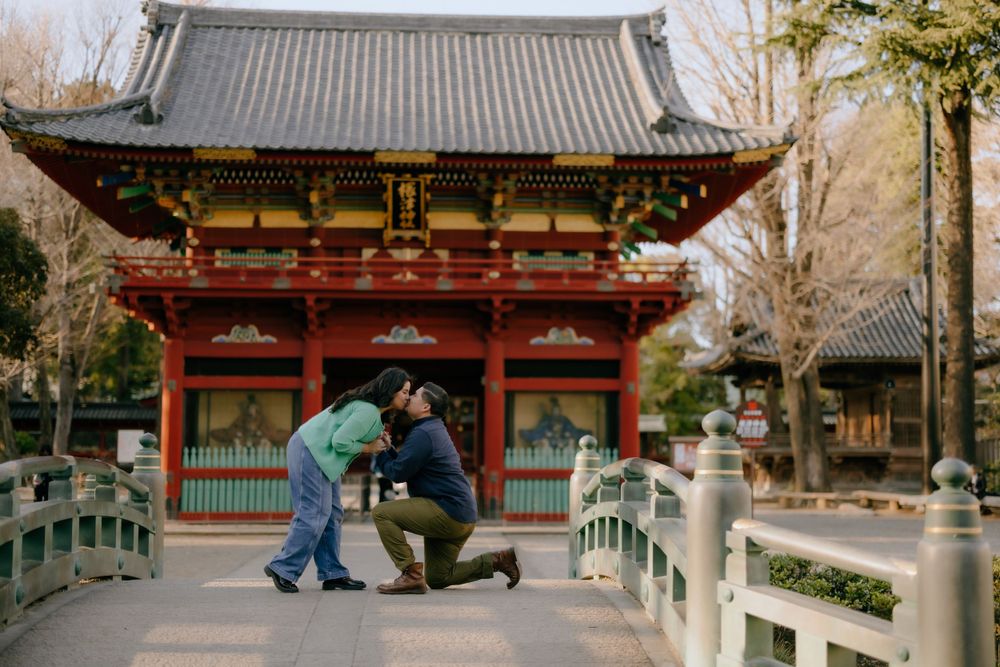 Two people in winter coats interact on a bridge in front of a traditional Japanese temple gate on a winter day.