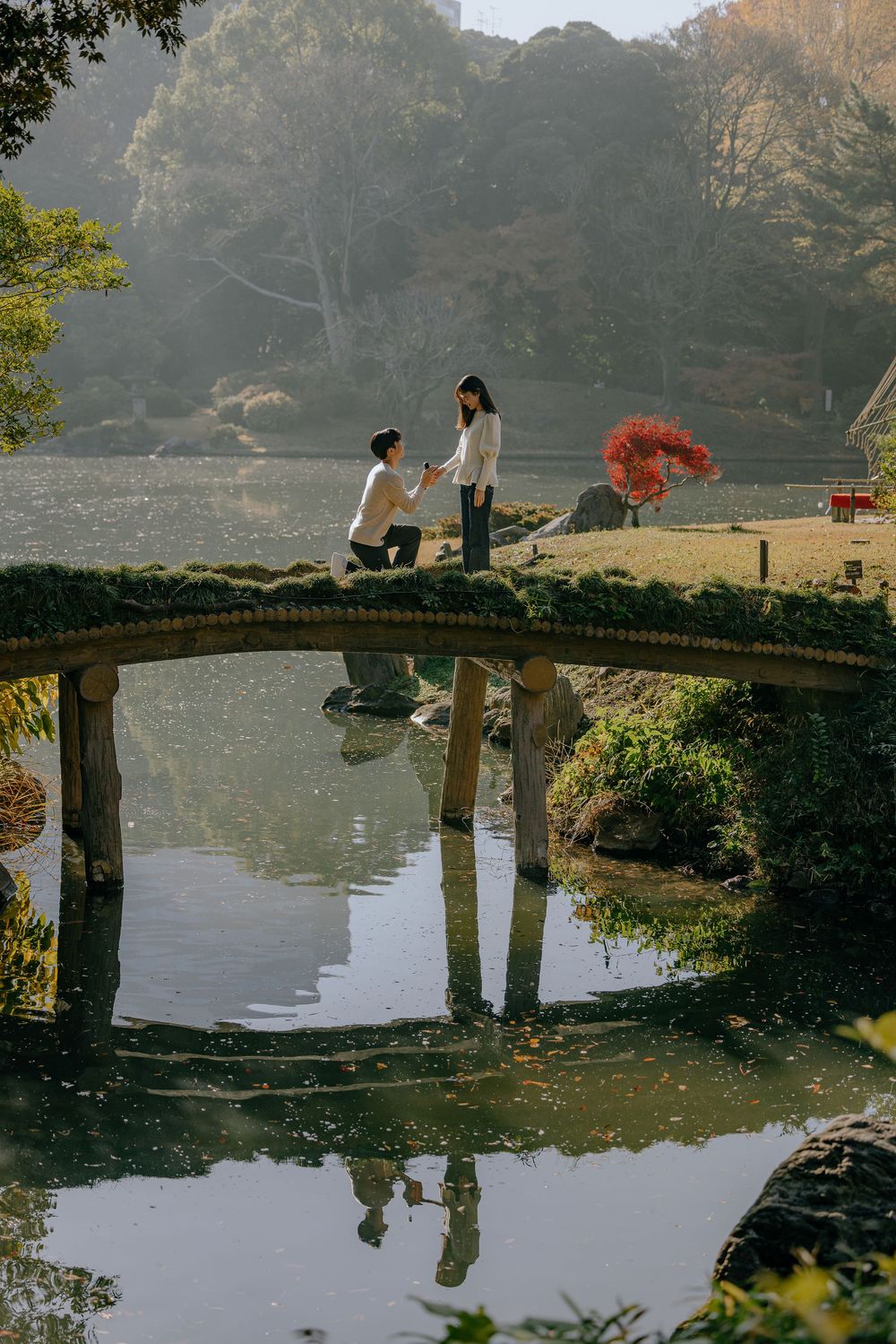 A serene pond reflects people sitting on a curved stone bridge in a tranquil Japanese garden during autumn.
