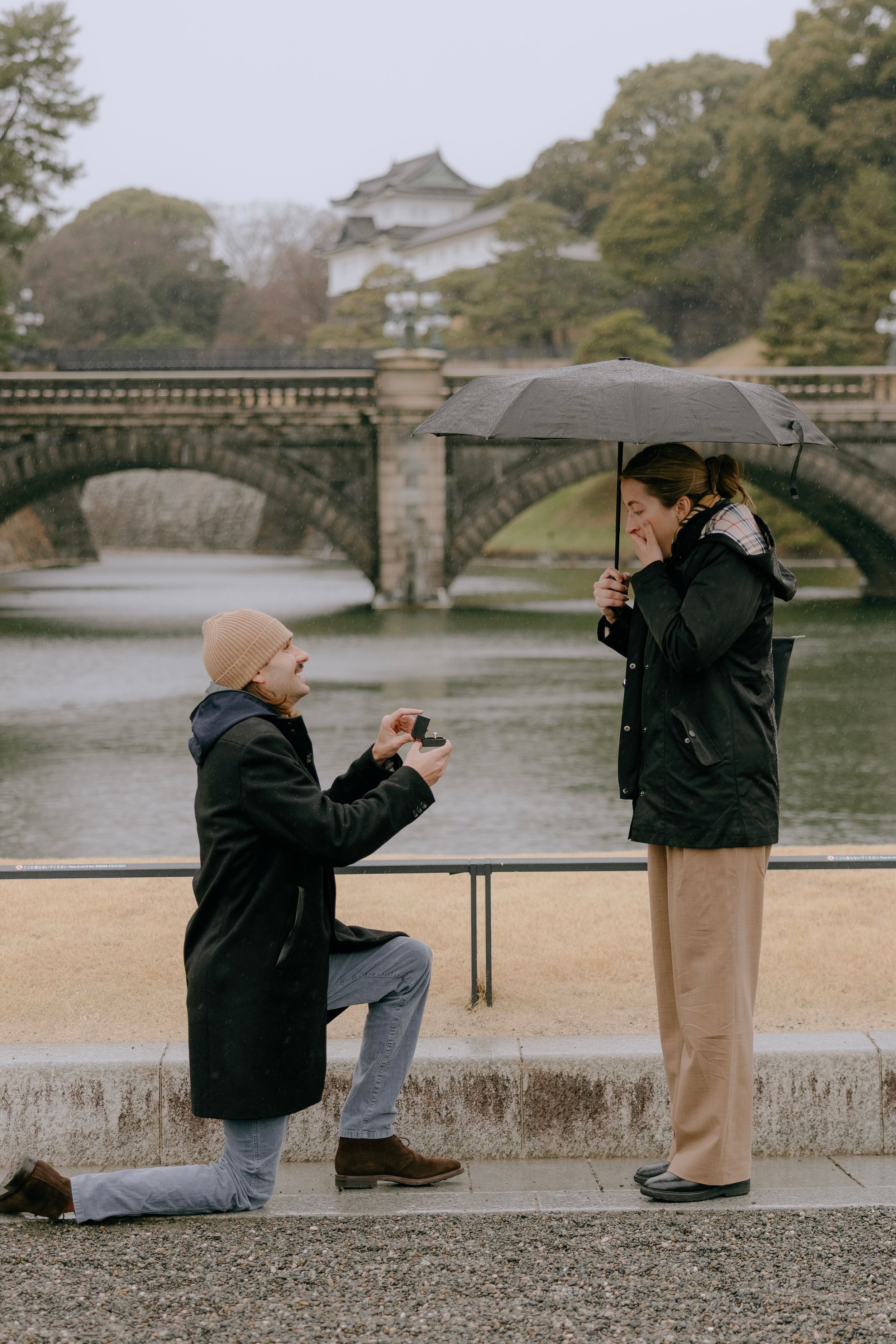 A romantic proposal moment near a historic stone bridge at the Imperial Palace in Tokyo, Japan on an overcast day.