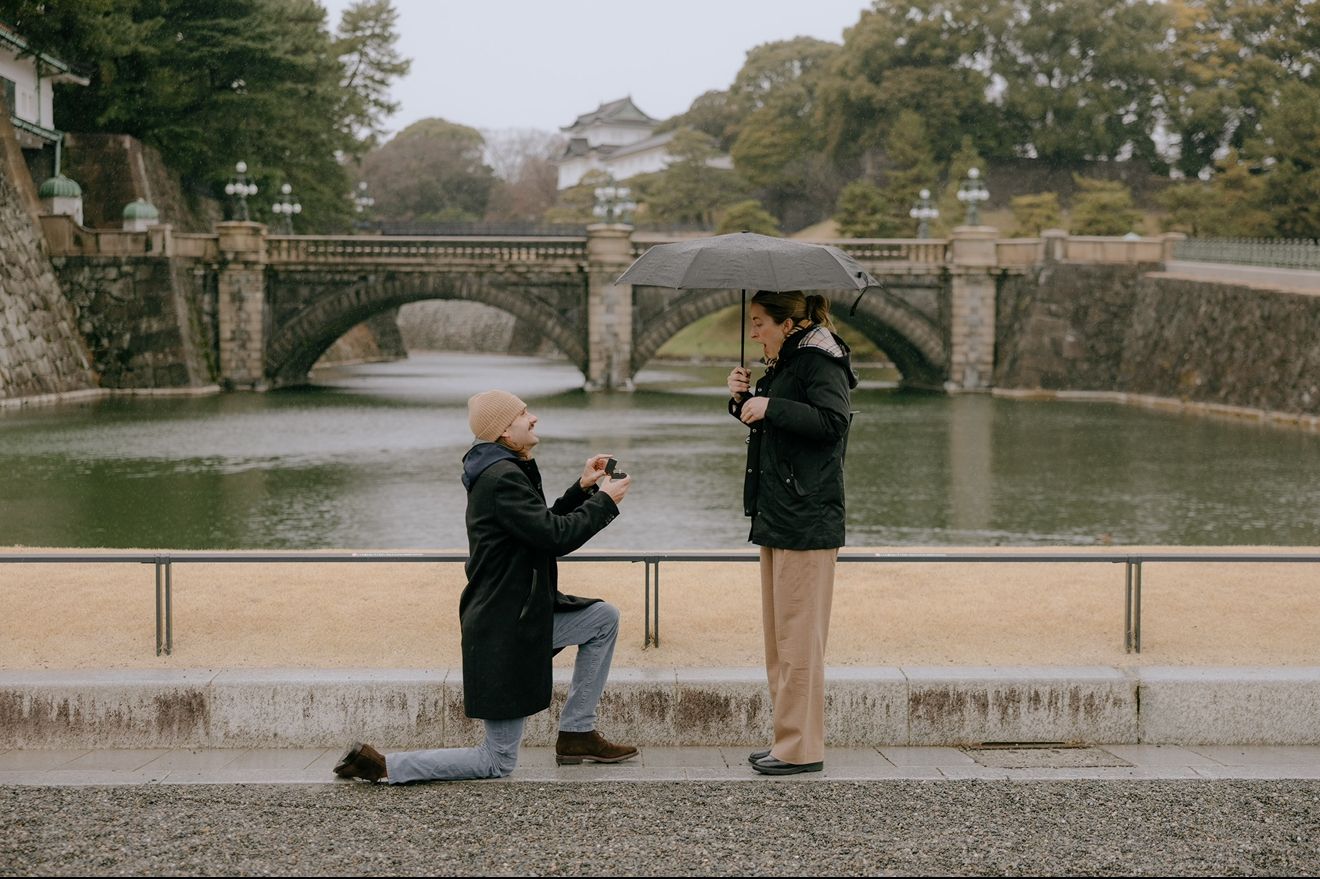 A proposal scene captured at an ornate stone bridge in Imperial Palace Gardens, Tokyo during winter.