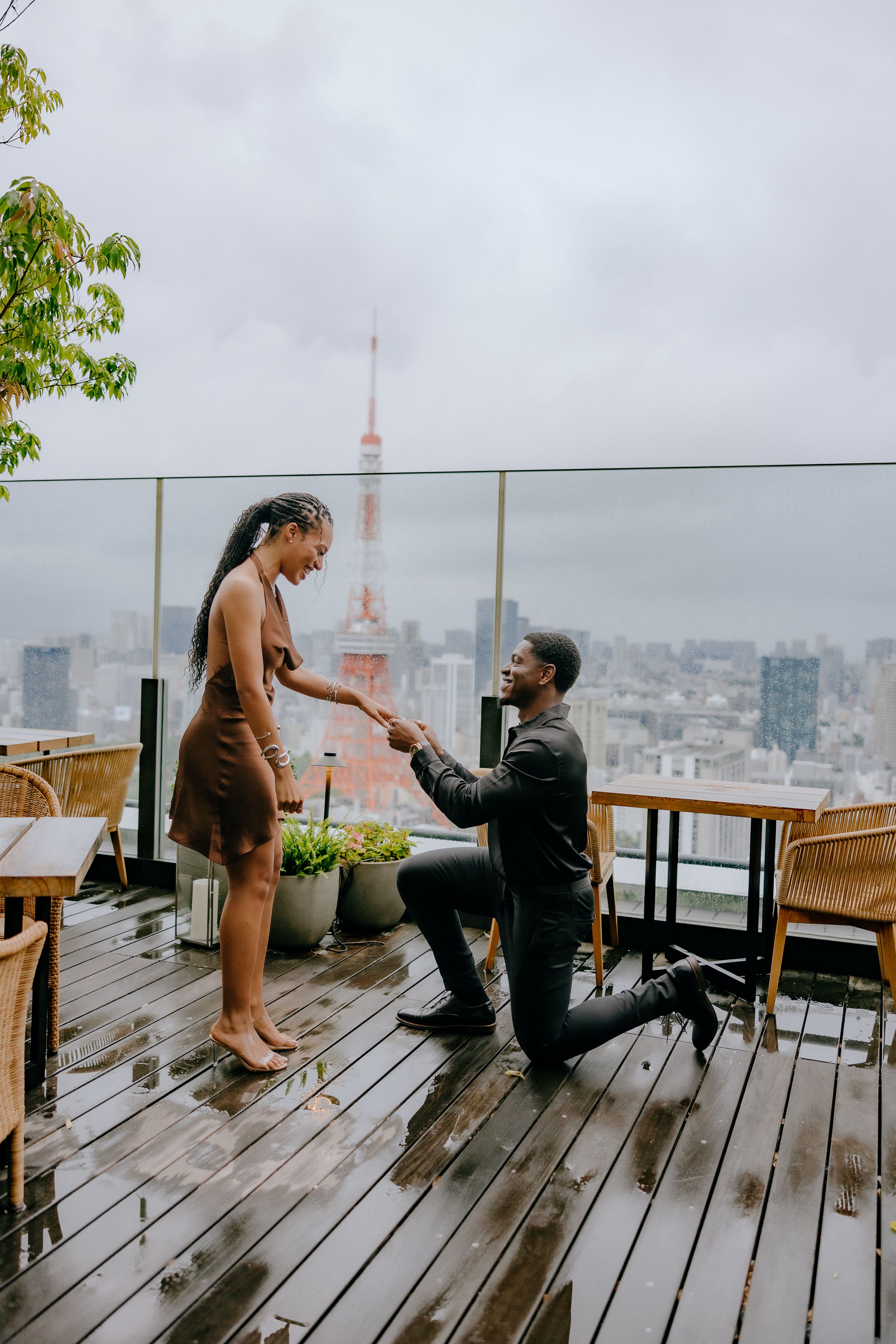 A couple shares a romantic moment during a rainy proposal on an outdoor deck overlooking a cityscape.