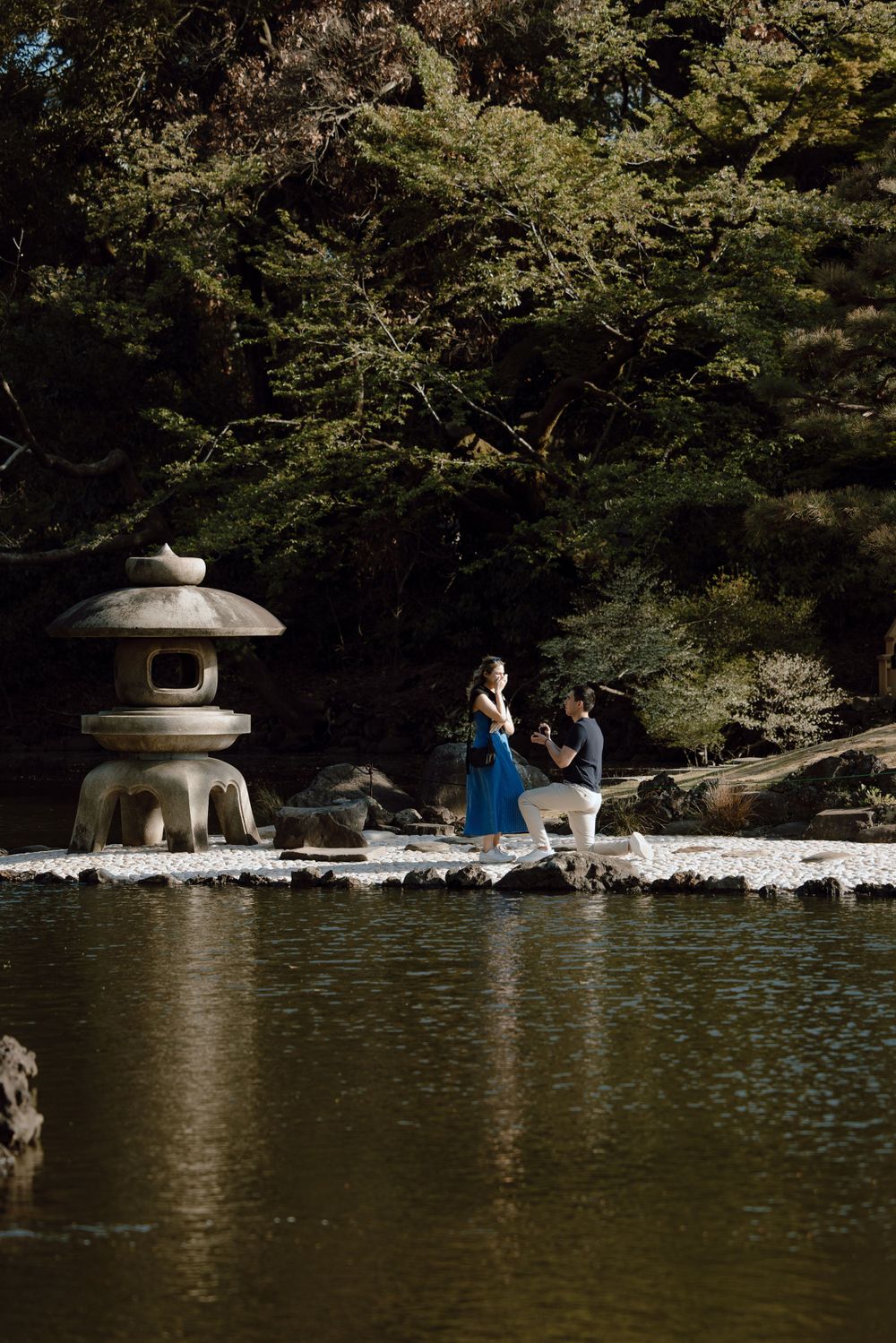A tranquil moment at a Japanese garden pond with a traditional stone lantern and two people walking along the water's edge.