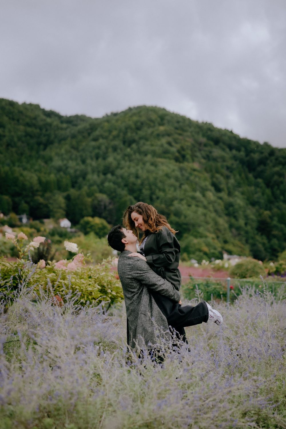 A couple embraces romantically in a field of purple wildflowers with green mountains in the background.
