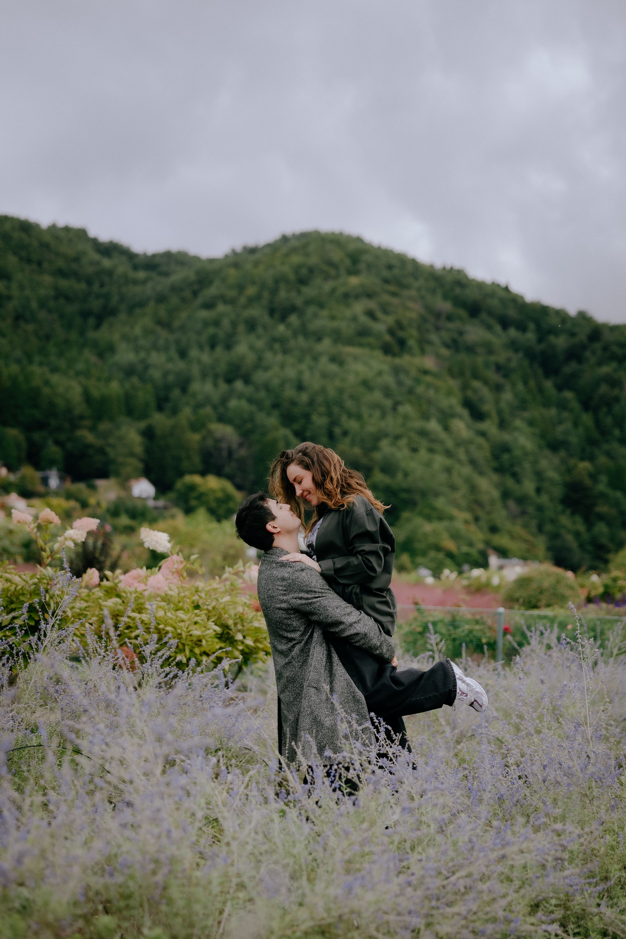 A couple embraces romantically in a field of purple wildflowers with green mountains in the background.