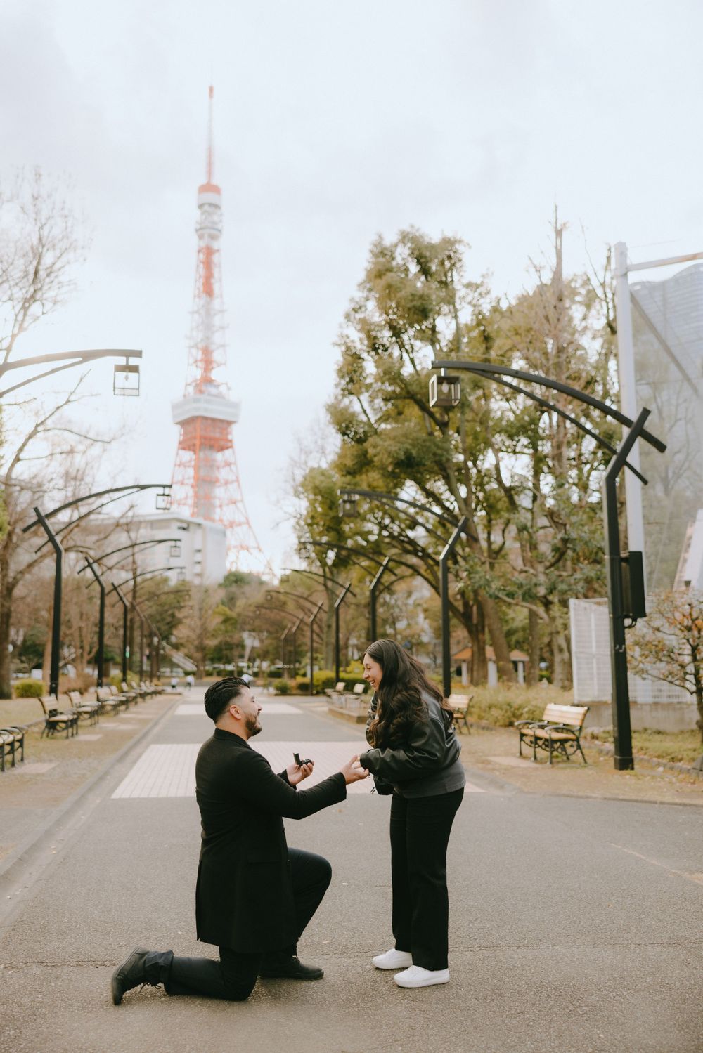 A romantic proposal scene on a path with Tokyo Tower rising in the background on a cloudy day.